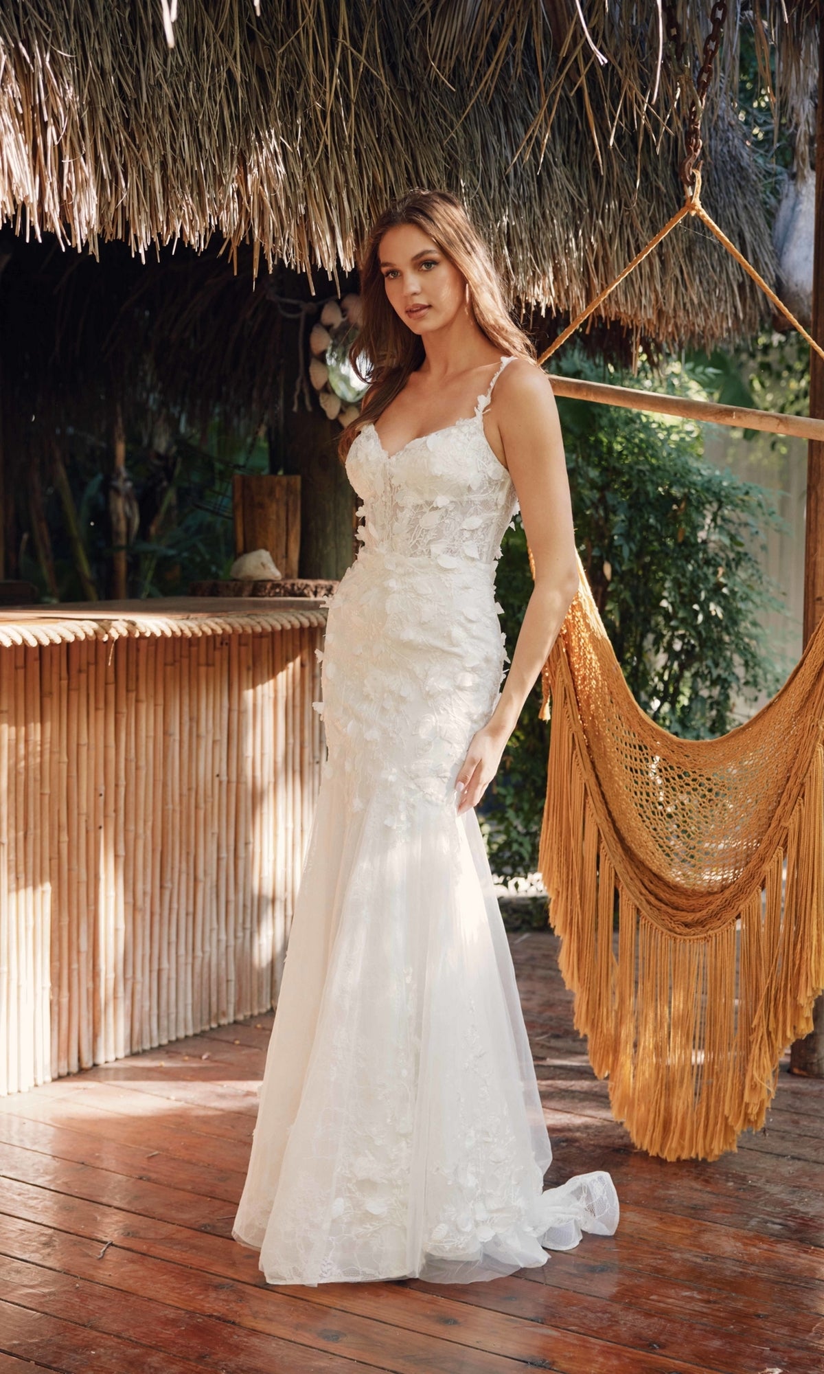 A woman in the Long Prom Dress JT2475UW by Juliet, an off-white mermaid gown with floral appliqué, stands on a wooden deck near a bamboo bar and mustard yellow hammock chair surrounded by greenery.
